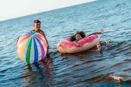 happy african american siblings having fun with beach ball and swimming tube in seaの写真素材