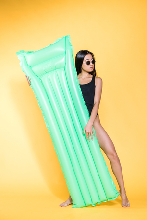 Full-length of beautiful asian woman in black swimsuit and sunglasses posing with a green pool mattress on yellowの写真素材