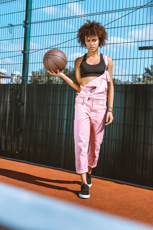 young african-american woman in sports bra and pink overalls holding basketball ball in her handの写真素材