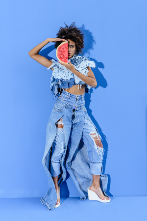 stylish african american model posing in denim clothes with slice of watermelon on blue の写真素材