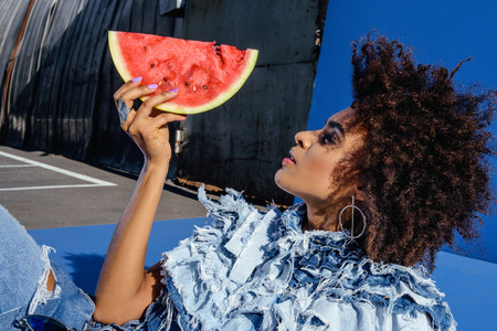 stylish afro girl posing in denim clothes with slice of watermelon  の写真素材