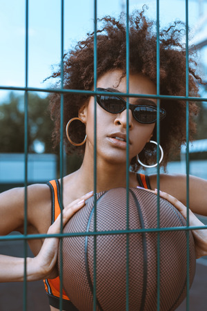 young african-american woman in sports bra and sunglasses holding a basketball ball in her hands in front of her chest, posing behind wired fencingの写真素材
