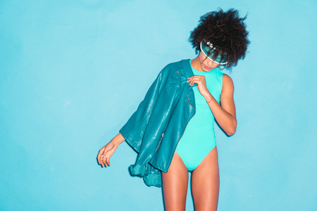 attractive stylish african american model posing in turquoise bodysuit and blue leather jacket,  isolated on turquoiseの写真素材