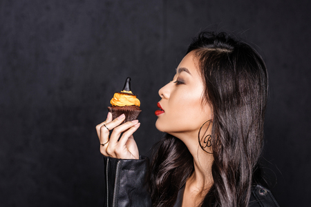 beautiful asian woman in leather jacket holding up a halloween cupcake and looking at itの写真素材
