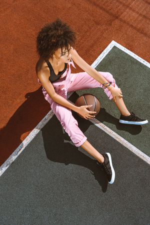 young african-american woman in sports bra and pink overalls sitting on ground of sports field with basketball ballの写真素材