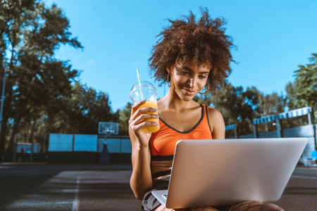 young african-american woman in bright sports bra sitting at sport court with laptop and orange juiceの写真素材