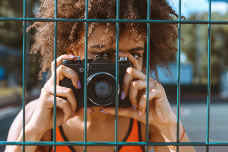 young african-american woman standing behind wired fencing and taking a picture with cameraの写真素材