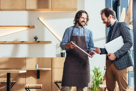 smiling bearded businessman and waiter standing and talking in cafeの写真素材