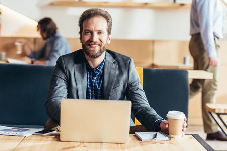handsome bearded man using laptop and smiling at camera while drinking coffee in cafeの写真素材