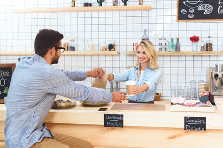 beautiful young waitress giving paper cup and take away food to client in cafeの写真素材
