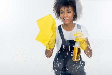 portrait of smiling african american cleaner in rubber gloves cleaning window isolated on whiteの写真素材