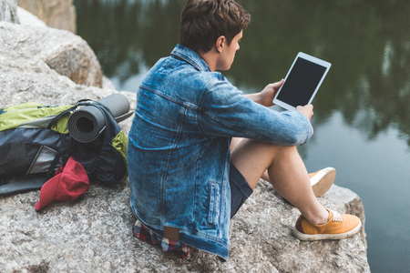 young man with backpack using tablet on nature while sitting on rocky river shoreの写真素材