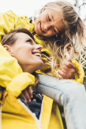 close up of cheerful mother piggybacking her daughter in yellow raincoat  の写真素材