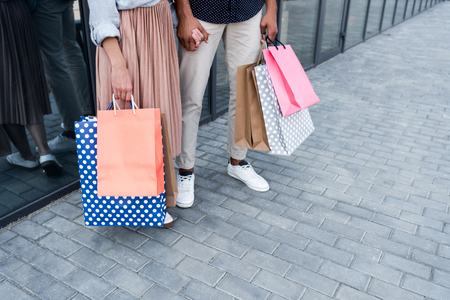 low section of young couple holding hands while standing with shopping bags on streetの写真素材