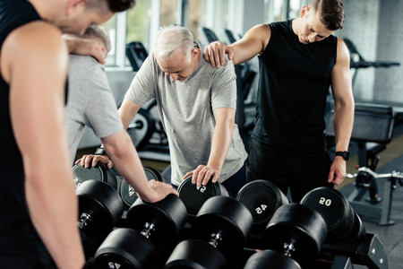 tired senior sportsman training with dumbbells and trainer in gym    の写真素材