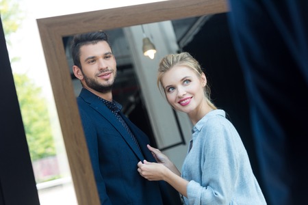 smiling young couple looking at mirror while choosing suit in boutiqueの写真素材