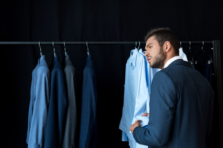 handsome young businessman looking away while choosing suit in boutiqueの写真素材