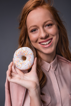 happy redhead elegant girl in pink clothes with sweet donut, isolated on grey の写真素材