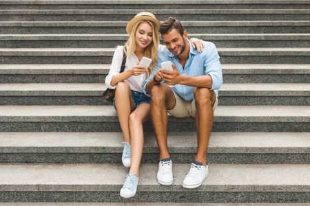 cheerful couple sitting on stairs and using smartphones on street