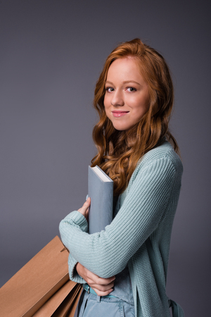 attractive elegant redhead girl in blue clothes with book and shopping bags, isolated on grey の写真素材