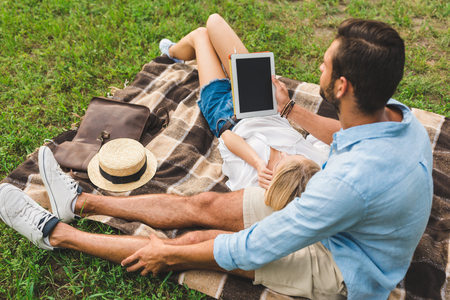 back view of young couple with tablet resting on blanket together in parkの写真素材