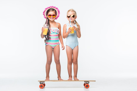 adorable little girls in sunglasses and swimsuits drinking orange juice while standing on skateboardの写真素材