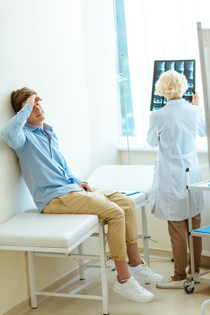 Young man sitting on a hospital daybed leaning on wall and touching his forehead with doctor examining his x-ray photographの写真素材