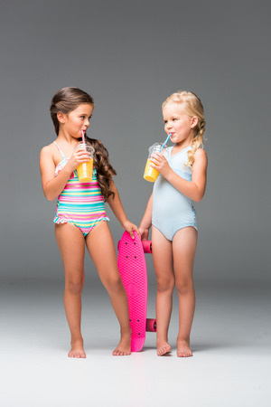cute little girls in swimsuits drinking orange juice while standing with skateboard isolated on greyの写真素材