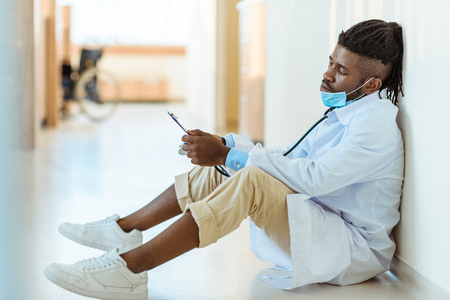 Young african-american doctor in lab coat sitting on floor in hospital corridor reading notes from clipboardの写真素材