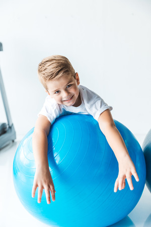 adorable little boy lying on fitness ball and smiling at camera on whiteの写真素材