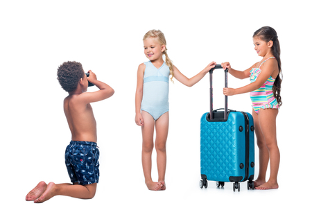 african american boy kneeling and photographing happy little girls with suitcase isolated on whiteの写真素材