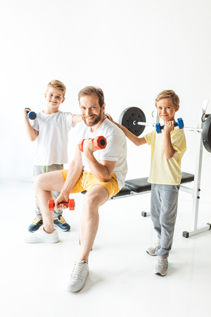 sportive father and sons exercising with dumbbells and smiling at camera on white の写真素材