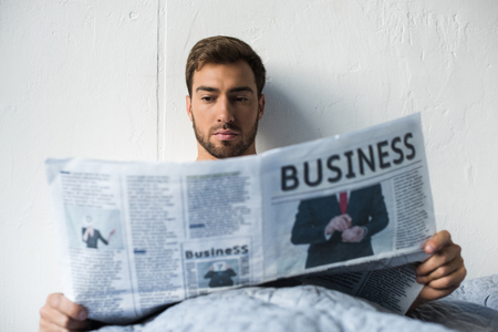 Attractive young man sitting in his bed and reading a business newspaperの写真素材