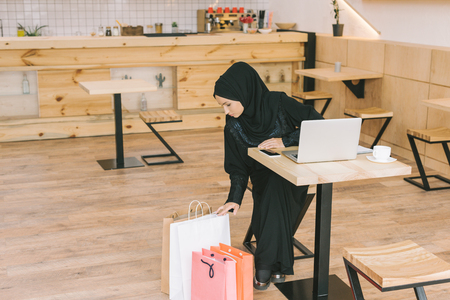 young muslim woman looking at shopping bags in cafe while sitting with laptopの写真素材