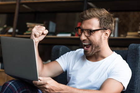 Young man sitting with laptop in armchair and cheering with his fist as he is looking at computer screenの写真素材