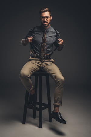 Handsome young man in glasses sitting on bar stool and pulling his suspendersの写真素材