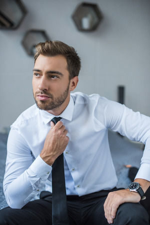 Young businessman in white shirt, straightening his tie and looking asideの写真素材