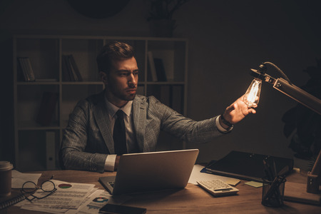 young businessman touching table lamp at workplace on late nightの写真素材