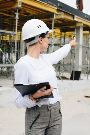 Young engineer in hardhat with clipboard at construction site, pointing her finger at unfinished buildingの写真素材