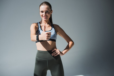 beautiful young sportswoman showing thumb up and smiling at camera isolated on greyの写真素材