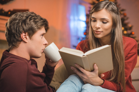 portrait of man with cup of hot drink looking at woman reading book at homeの写真素材