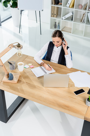 Young businesswoman sitting at desk in office, while talking on headset and writing in notebookの写真素材