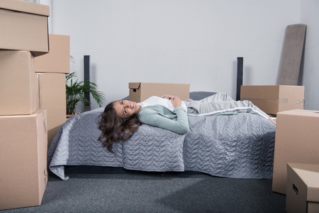 tired woman lying on bed after unpacking cardboard boxes at new home, moving home conceptの写真素材