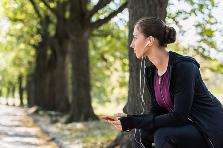 thoughtful woman listening music in park while sitting on benchの写真素材