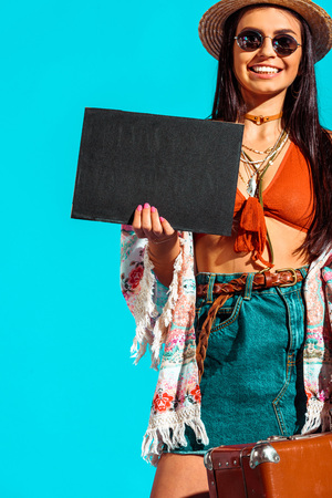 attractive female bohemian traveler with black board and suitcase isolated on turquoise  の写真素材
