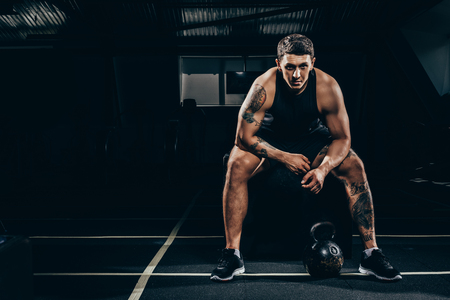 Athletic sportsman sitting in gym and looking at camera with kettlebell placed on floorの写真素材