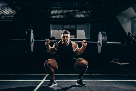 Athletic sportsman squatting while lifting a barbell with weights at gym and looking at cameraの写真素材