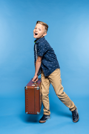 cheerful little boy holding suitcase and looking at camera isolated on blueの写真素材