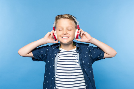 happy little boy with closed eyes listening music in headphones isolated on blue