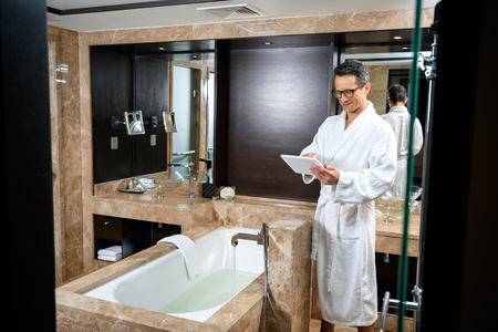 Businessman wearing a bathrobe in hotel bathroom, using his digital tablet while standing over bathtubの写真素材